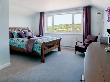 A bedroom with wooden bed and purple bedding next to a window with curtains and a chair at Carousel in Broad Haven