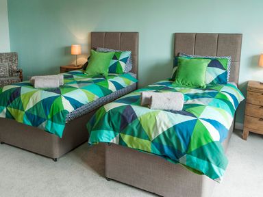 A bedroom with two single beds with multicolored bedding green pillows lamps on bedside tables and a patterned armchair at Carousel in Broad Haven
