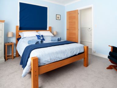 A bedroom with a wooden bed frame two rolled towels bedside tables with lamps and a door leading to a bathroom at Carousel in Broad Haven