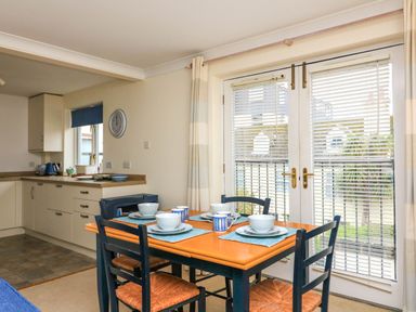 A dining area with a wooden table set with bowls and mugs and four chairs next to glass doors with blinds at 8 Dartmouth House in Dartmouth