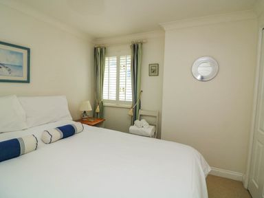 A bedroom with a bed two bolster pillows a window with curtains a side table with lamp a chair with towels and wall decorations at 8 Dartmouth House in Dartmouth