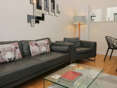 A living room with a black leather sofa and chair with sheep cushions a glass coffee table wooden floor and a floor lamp at 12 Blue Hill Park in Ambleside