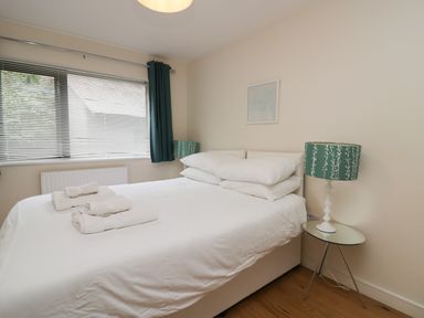 A bedroom with a white bed and folded towels on top beside a window with blinds and green curtains at 12 Blue Hill Park in Ambleside