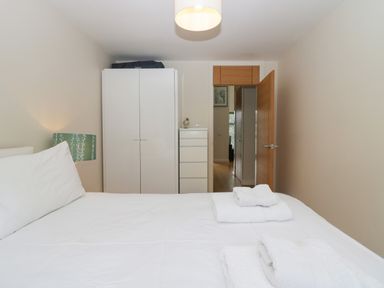 A bedroom with a bed white bedding folded towels a white wardrobe a chest of drawers a green bedside lamp and an open wooden door at 12 Blue Hill Park Ambleside