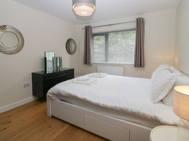 A bedroom with a white bed and folded towels on it next to a black dresser with mirrors and windows with curtains at 12 Blue Hill Park in Ambleside