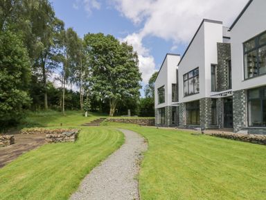 A pathway through a lawn leading to modern white houses with large windows surrounded by trees at 12 Blue Hill Park in Ambleside