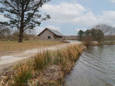 A wooden cabin near a dirt path and a body of water with trees in the background at Kingfisher Lodge in Treverbyn near St Austell