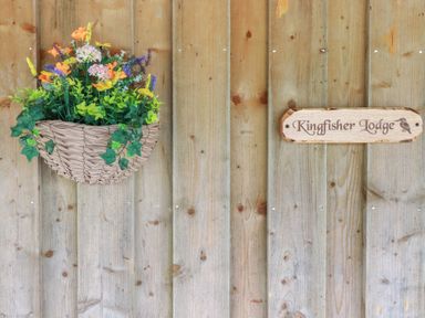 A wooden wall with a hanging basket of flowers and a sign reading Kingfisher Lodge in Treverbyn near St Austell