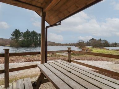 A wooden picnic table and bench on a covered porch overlooking a lake with trees on the shore at Kingfisher Lodge in Treverbyn near St Austell
