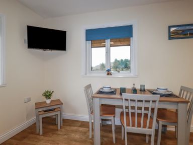 A dining area with a wooden table set for two and four chairs a small table with a plant a wall mounted television and a window with a blue blind at Kingfisher Lodge in Treverbyn near St Austell