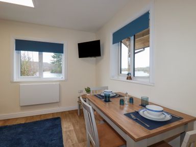 A dining area with a wooden table set for two near windows with blue blinds and a wall-mounted television at Kingfisher Lodge in Treverbyn near St Austell