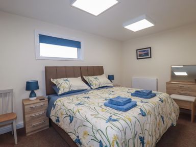 A bedroom with a double bed, bedside tables with lamps, a chair, a dresser with a mirror and a framed picture on the wall at Kingfisher Lodge Treverbyn near St Austell