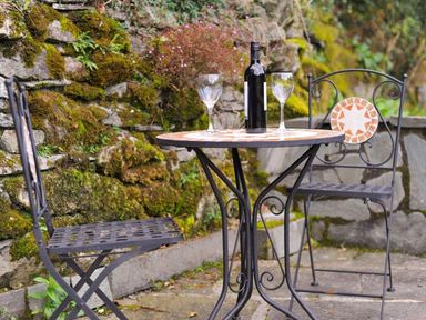 A small round table with a wine bottle and two wine glasses with two metal chairs outside near a mossy stone wall at Riverbank At Stepping Stones in Rydal
