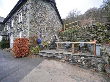 An exterior stone wall of a house with steps leading to a raised patio with two chairs and a small table at Riverbank At Stepping Stones in Rydal