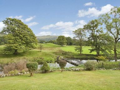 A grassy field with trees and a small river with stepping stones at Riverbank At Stepping Stones in Rydal