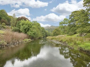 A river flowing between grassy banks with trees and hills in the background at Riverbank At Stepping Stones in Rydal