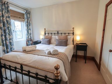 A bedroom with a metal-framed bed with pillows and rolled towels on it two bedside tables with lamps and a window with floral curtains at Riverbank At Stepping Stones in Rydal