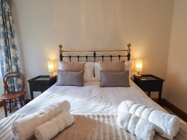A bedroom with a bed with pillows and folded towels bedside tables with lamps and a wooden chair with floral curtains at Riverbank At Stepping Stones in Rydal