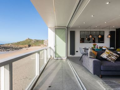 A balcony overlooking a sandy beach and rocky hill with a living room and dining area visible through open glass doors at Louies Loft in St Ives
