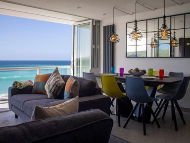 A living room with a gray sofa and colorful pillows next to a dining table with multicolored chairs and ocean view at Louies Loft in St Ives