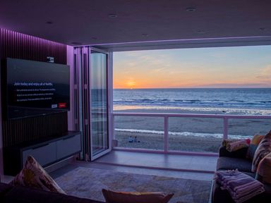 A living room with a television a sofa and an open wall showing a beach and ocean view at Louies Loft in St Ives