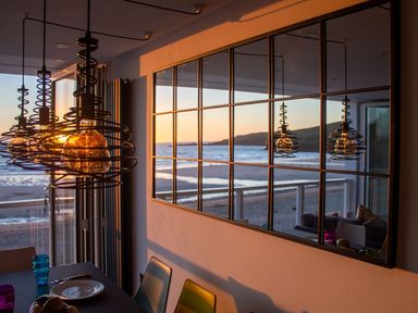 A dining area with hanging light fixtures and a large window reflecting the beach at Louies Loft in St Ives