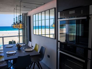 A dining area with a table set with plates and glasses next to a large window overlooking a beach at Louies Loft in St Ives