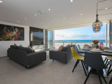 A living room with dark sofas and a dining table with colorful chairs facing a beach view at Louies Loft in St Ives