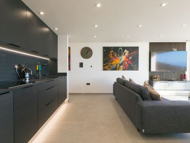 A kitchen with black cabinets and appliances next to a living area with a gray sofa and wall-mounted TV at Louies Loft in St Ives