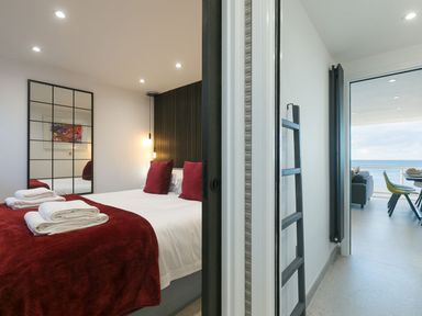 A bedroom with a bed and towels beside an open door leading to a living area with chairs and an ocean view at Louies Loft in St Ives
