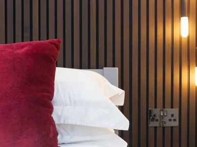 A bed with white pillows and a red cushion against a dark wood slatted wall with mounted lights and power outlets at Louies Loft in St Ives