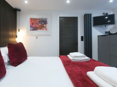 A bedroom with a bed with red cushions and white sheets a dark wooden door a wall-mounted tv and a painting at Louies Loft in St Ives