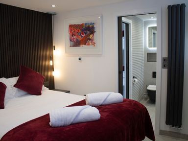 A bedroom with a bed with white sheets and red pillows and blanket with two rolled white towels on the bed and a bathroom visible through an open door at Louies Loft in St Ives