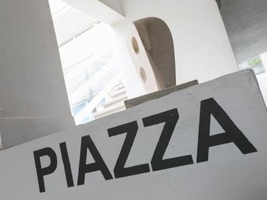 A white wall with the word PLAZZA in black letters and a stone sculpture behind it