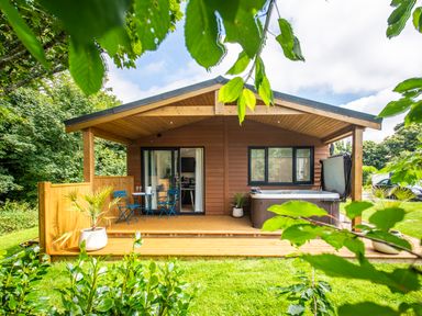 A wooden cabin with a small porch table and chairs a hot tub and greenery around at Maple Lodge in Angarrack