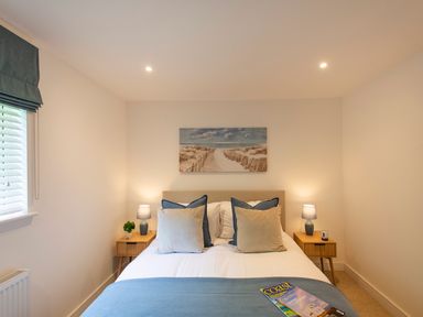 A bedroom with a bed between two wooden nightstands with lamps and a seaside painting on the wall at Maple Lodge in Angarrack