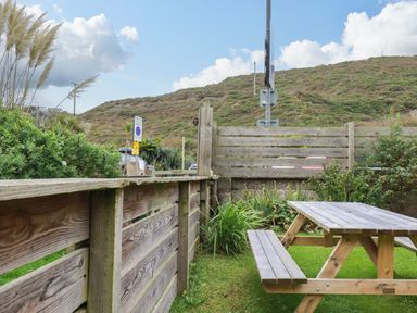 A wooden picnic table and bench on grass near a wooden fence with hills and road signs in the background at 1 Mill Cottages in Porthtowan