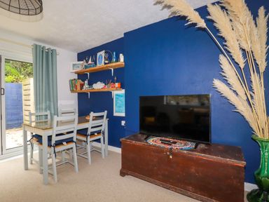 A dining table with four chairs near sliding glass door and TV on wooden chest against blue wall at 1 Mill Cottages in Porthtowan