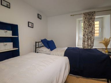 A bedroom with two single beds one with blue pillows and blanket a window with floral curtain and shelves with white baskets at 1 Mill Cottages in Porthtowan
