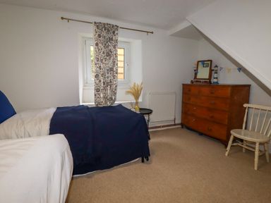 A bedroom with two single beds a floral curtain on the window a wooden dresser and a wooden chair at 1 Mill Cottages in Porthtowan