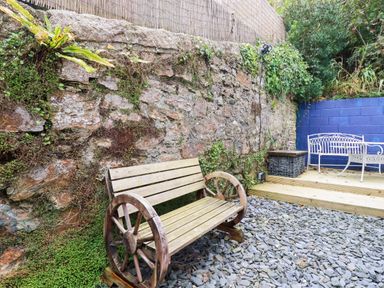 A garden area with a wooden bench with wagon wheels against a stone wall and a raised deck with white metal bench at 1 Mill Cottages Porthtowan