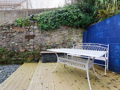 An outdoor seating area with a white metal table and bench on a wooden deck next to stone and blue painted walls with greenery at 1 Mill Cottages in Porthtowan