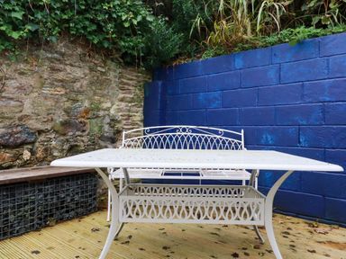 An outdoor seating area with a white metal table and bench against a stone wall and blue painted brick wall at 1 Mill Cottages in Porthtowan