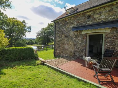 A stone house with a wooden deck and chairs near a grassy yard and pond at The Old Workshop in Dre-Fach near Lampeter