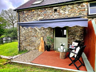 An outdoor patio area with a stone wall house a green door a glass table on a wooden base two black chairs with cushions and a retractable awning at The Old Workshop in Dre-Fach near Lampeter