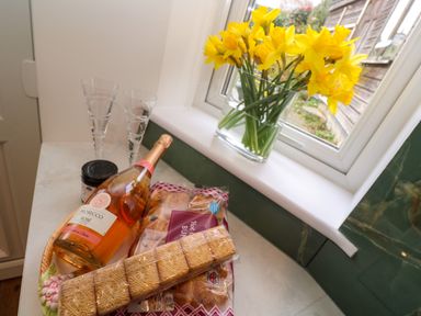 A kitchen counter with a bottle of prosecco rose glassware biscuits hot cross buns and a vase of yellow flowers at Elder Cottage Studio in Perranwell Station near Ponsanooth
