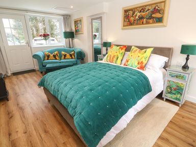A bedroom with a bed a teal comforter colorful pillows a teal sofa lamps and paintings at Elder Cottage Studio in Perranwell Station near Ponsanooth