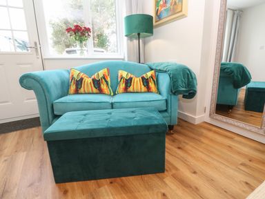 A teal sofa with colorful pillows and a matching ottoman in a room with a wooden floor and a large mirror at Elder Cottage Studio Perranwell Station near Ponsanooth
