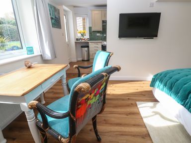 A room with two blue upholstered chairs at a wooden table near a window a wall-mounted TV and a bed with a blue blanket at Elder Cottage Studio in Perranwell Station near Ponsanooth