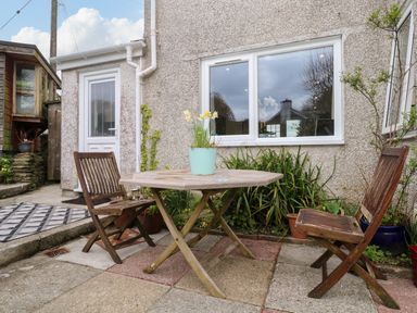 An outdoor patio with a wooden table, two wooden chairs, potted plants, and a white door on a pebble dash wall at Elder Cottage Studio in Perranwell Station near Ponsanooth